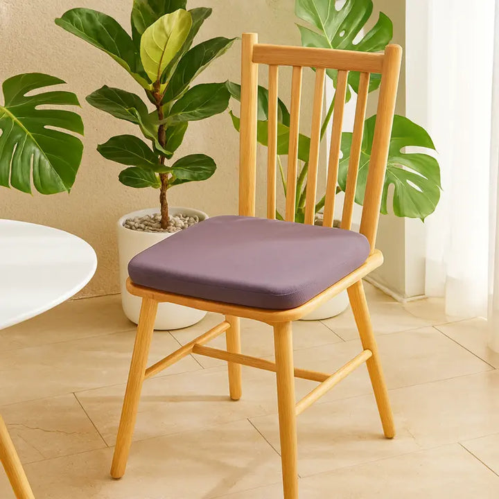 Une chaise en bois clair avec un coussin de siège carré violet à mémoire de forme est placée sur une terrasse lumineuse et minimaliste.