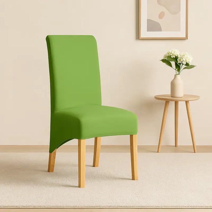 Chaise recouverte d’une housse de couleur verte, présentée dans un intérieur minimaliste et lumineux. La chaise est placée sur un tapis beige, devant un mur clair décoré d’un tableau et d’un pot de fleurs sur une table en bois clair. L’éclairage naturel met en valeur la texture douce et élégante du tissu, créant une ambiance chic et raffinée, idéale pour une présentation produit.