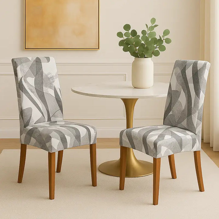 Deux chaises élégantes recouvertes de housses à motifs gris et blancs entourent une table ronde au pied doré et plateau blanc, posée sur un tapis beige. Un grand vase blanc orné de branches vertes décore la table. Le fond présente un mur crème à moulures et un tableau abstrait dans les tons dorés, créant une atmosphère chic, épurée et lumineuse.