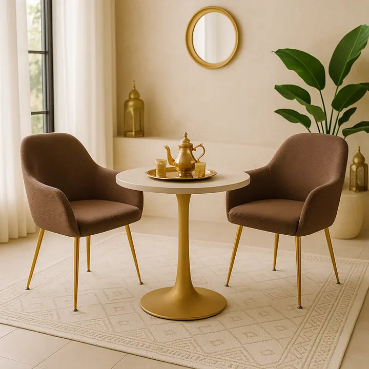 Deux chaises recouvertes de housses en jacquard marron avec pieds dorés, disposées autour d’une table ronde dorée dans un petit salon de thé luxueux. Un service à thé marocain doré est posé sur la table, entouré de lanternes et de plantes vertes, dans une ambiance lumineuse et élégante aux tons beiges et dorés.