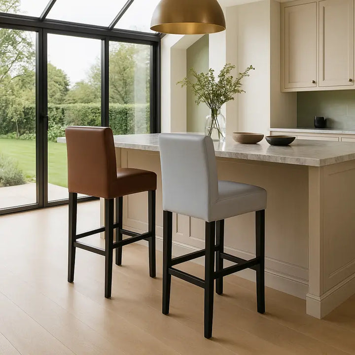 Deux chaises de bar avec housses en simili cuir imperméable, l’une blanche et l’autre marron, disposées autour d’un îlot central dans une cuisine moderne et luxueuse. L’espace est lumineux, avec un plan de travail en marbre beige, un grand luminaire doré, un sol en bois clair et une baie vitrée donnant sur un jardin verdoyant.
