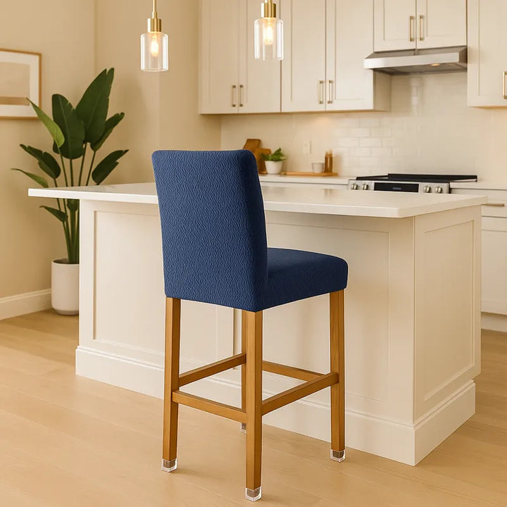 Cuisine américaine moderne et lumineuse avec un îlot central beige et en démo, une chaise de bar recouverte d'une housse en jacquard bleu marine. Le décor comprend des meubles en bois clair, des plantes vertes et des suspensions dorées, créant une ambiance élégante, chaleureuse et contemporaine.