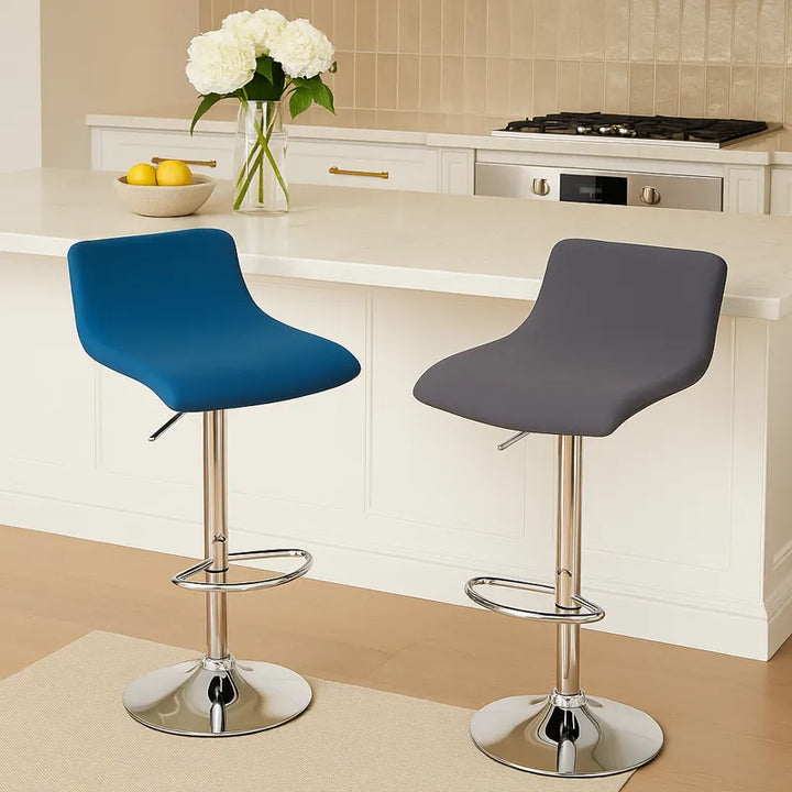 Deux chaises de bar rotatives, l’une bleue et l’autre grise, sont installées devant un comptoir en marbre clair dans une cuisine moderne et luxueuse. Le décor est lumineux, agrémenté d’un vase de fleurs blanches, d’un bol de citrons et de détails dorés, créant une atmosphère élégante et contemporaine.
