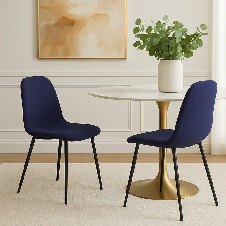 Deux chaises au design moderne, revêtues de tissu bleu marine, entourent une table ronde à pied doré et plateau blanc. Sur la table, un grand vase blanc contenant des branches vertes apporte une touche naturelle. En arrière-plan, un tableau aux tons beige et ocre orne un mur blanc à moulures, dans une pièce lumineuse et élégante.