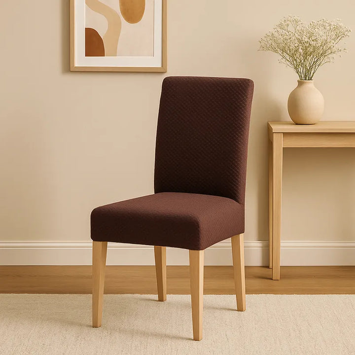 Chaise élégante recouverte d’une housse en jacquard effet tressé de couleur marron, placée dans un intérieur beige minimaliste et lumineux. En arrière-plan, un cadre décoratif abstrait et un petit vase en céramique beige avec des fleurs blanches posés sur une console en bois clair complètent l’ambiance chic et apaisante.