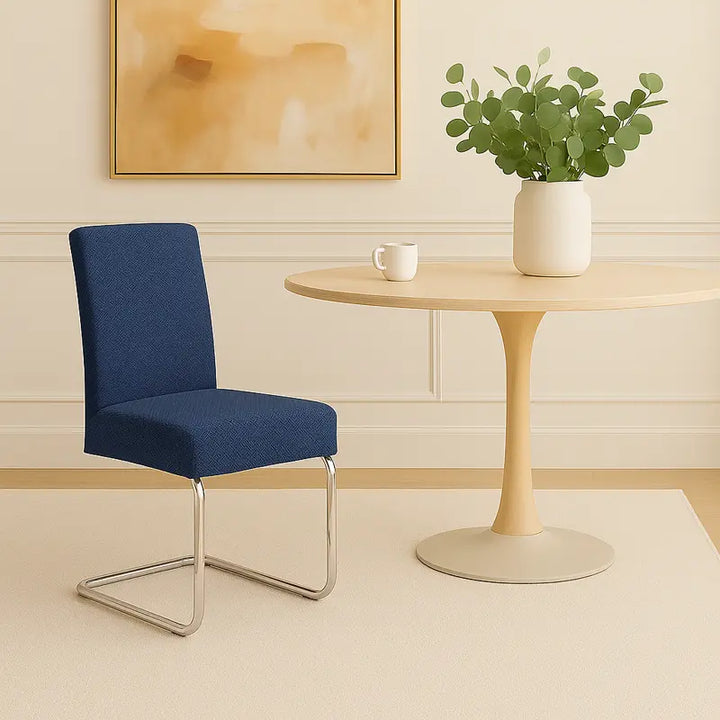 Une chaise moderne recouverte d’une housse en jacquard bleu effet tressé est placée à côté d’une table ronde en bois clair, sur un tapis beige. Un vase blanc contenant des branches vertes et une petite tasse décorent la table. L’arrière-plan présente un mur crème à moulures et un tableau abstrait beige, dans un décor minimaliste, lumineux et élégant.