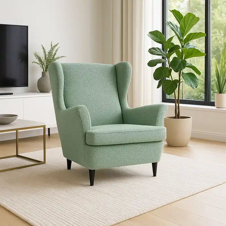 Salon contemporain luxueux et épuré, avec un fauteuil à oreilles recouvert d’une housse vert clair en jacquard effet tressé. La pièce baigne dans une lumière naturelle grâce à de grandes baies vitrées, décorée de plantes vertes et d’éléments modernes dans une ambiance élégante et apaisante.