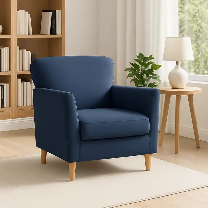 Un fauteuil club en velours bleu mis en valeur dans un salon minimaliste et lumineux, avec un tapis beige, une bibliothèque élégante.