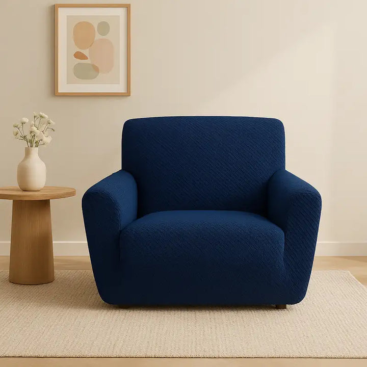 Fauteuil recouvert d’une housse bleu marine en jacquard effet tressé, mis en valeur dans un salon beige minimaliste et lumineux. L’ambiance est chic et élégante, avec un tapis clair, un tableau mural et un vase de fleurs blanches en arrière-plan, soulignant la texture tressée du tissu.