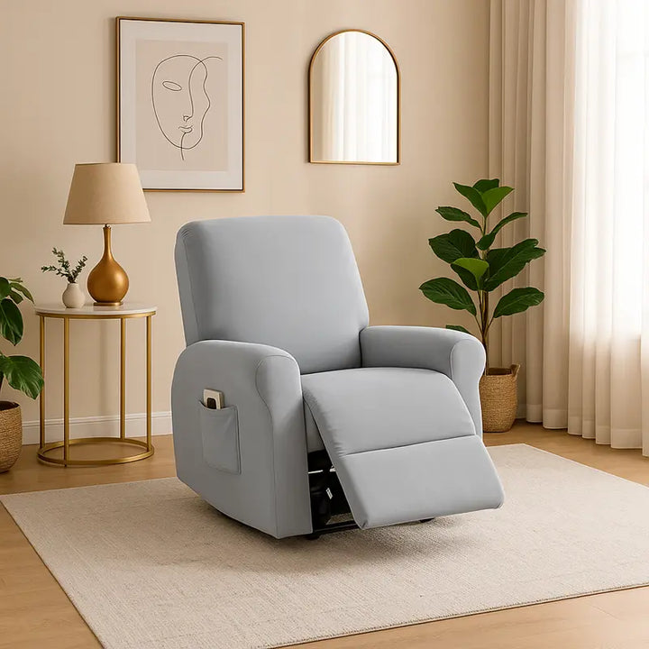 Un fauteuil inclinable gris clair recouvert d’une housse élégante est placé sur un tapis beige dans un salon lumineux et raffiné, entouré de plantes vertes, d’une table dorée avec lampe beige, d’un miroir doré et d’un tableau minimaliste, dans une ambiance apaisante et luxueuse