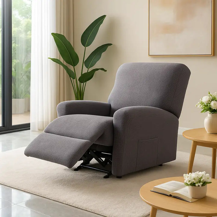 Un fauteuil relax inclinable recouvert d’une housse en jacquard gris imperméable, placé dans un salon lumineux et luxueux avec tapis moelleux, pot de fleurs et grande baie vitrée laissant entrer la lumière naturelle.
